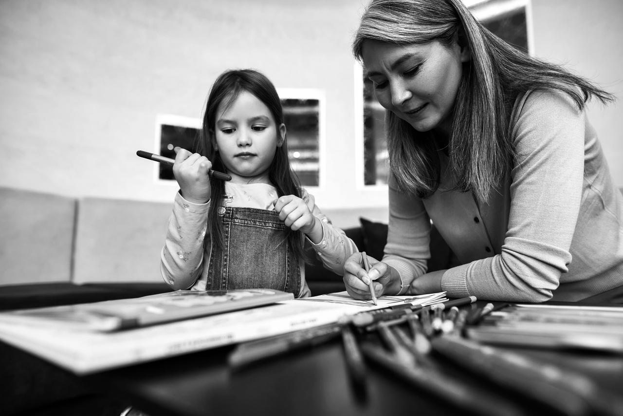 Little girl holding felt-tip pen, sitting at wooden table next to mature woman, drawing on sheet of paper