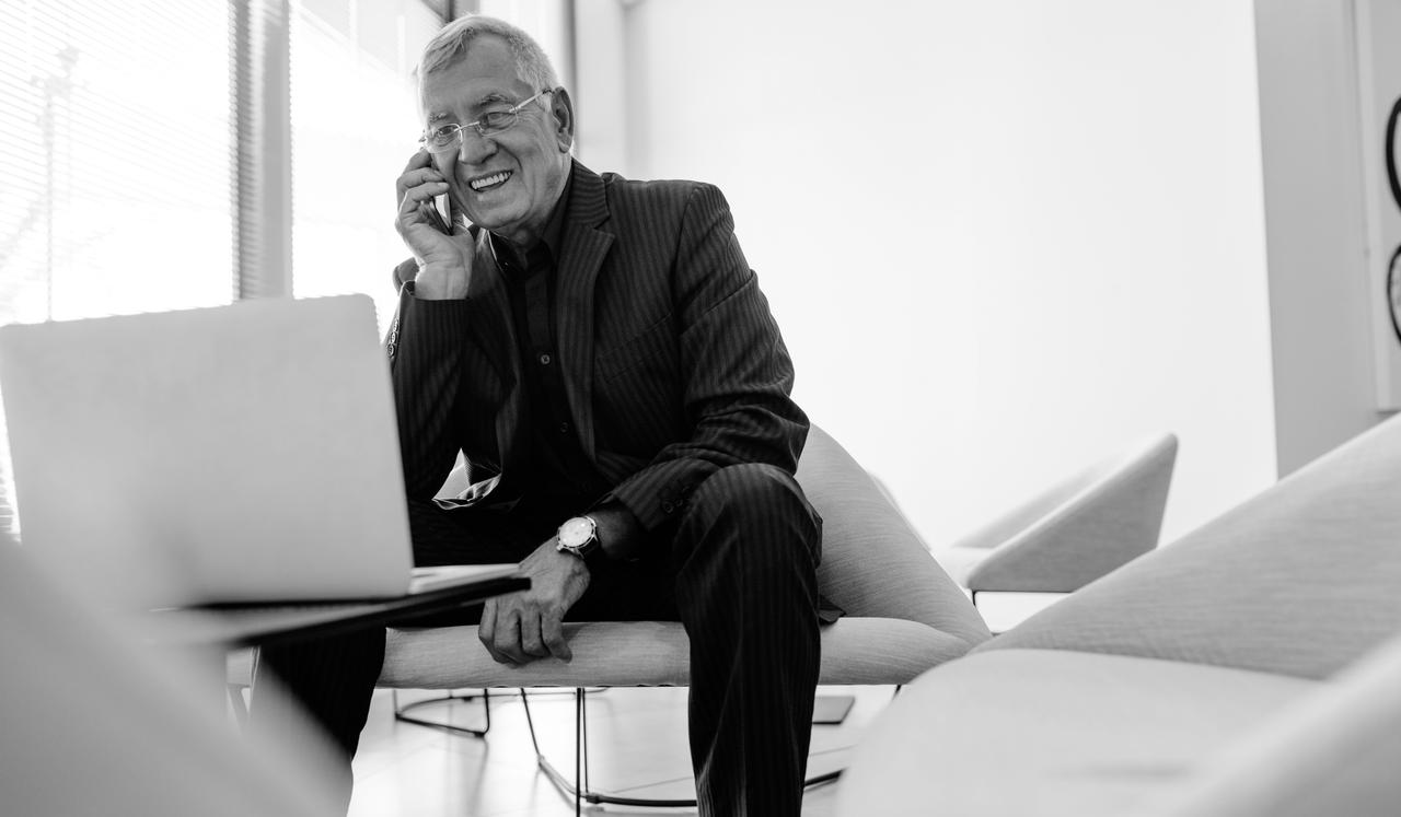 Mature businessman sitting in office lobby with a laptop using mobile phone.